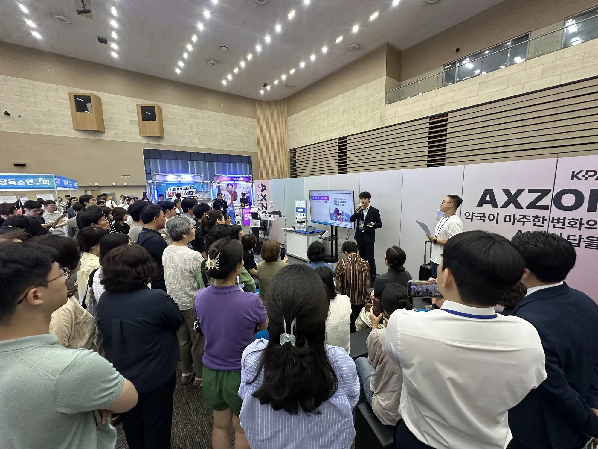 광주 팜엑스포 행사장에서 마이크를 들고 발표하는 강재현 약사와, 서거나 앉아 이를 지켜보는 많은 참관객.